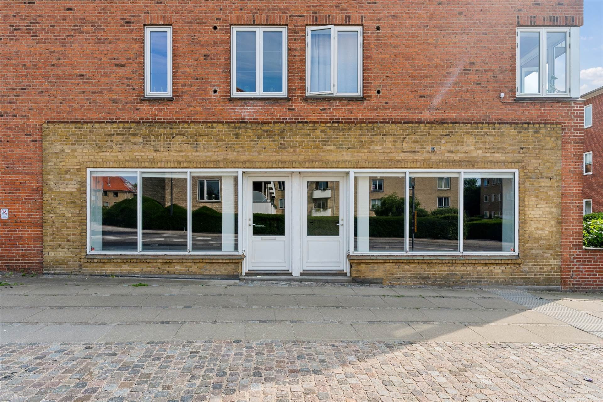 Et lejemål med glasvinduer og hvide døre foran en rødmalet murstensbygning med fem hvide vinduer. En træbrolægning ud for