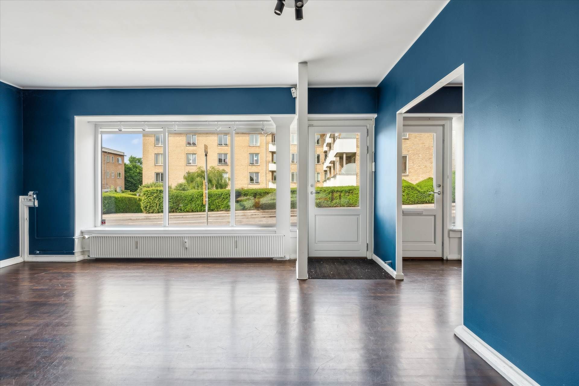 Et stort værelse med høje lofter, gulvbelægning, en hvid dør og en stor vinduesfront med udsigt til grønne områder udenfor