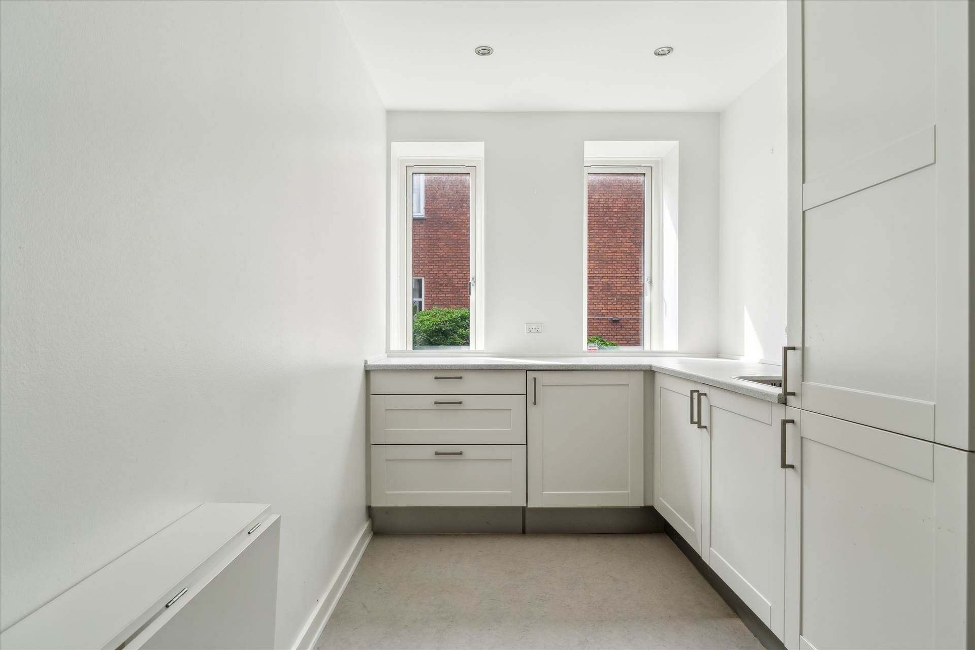 Lyst og rummeligt køkken med hvide vægge, køkkenskabe, komfur og vindue til gaden