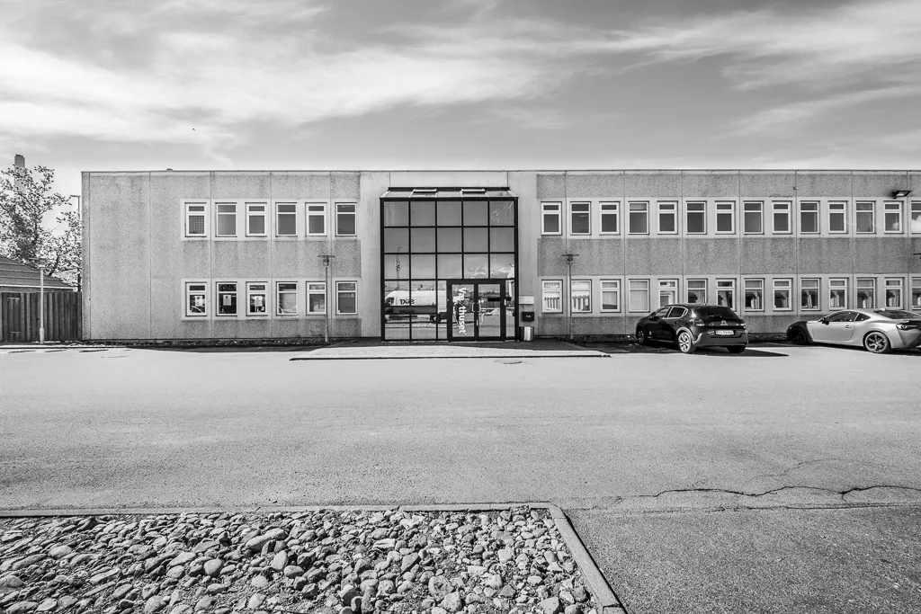 Et moderne kontorbygning med glasdøre og flere vinduer, to biler parkeret foran og en udsigt til en motorvej og busser i