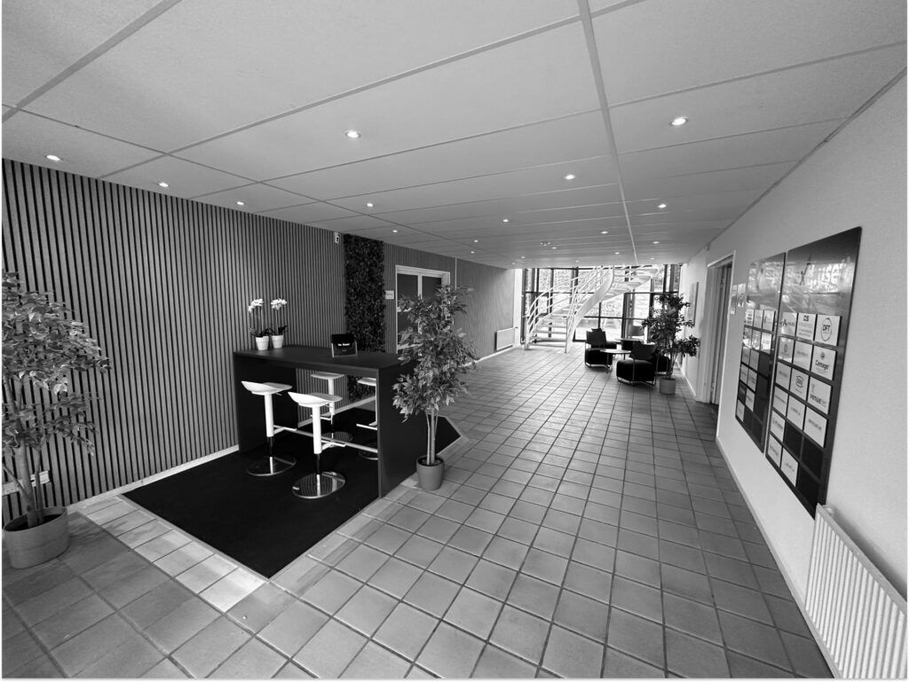 Hall med lyse cellekontorer, to stolpe og tre potteplanter samt træstiger i baggrunden