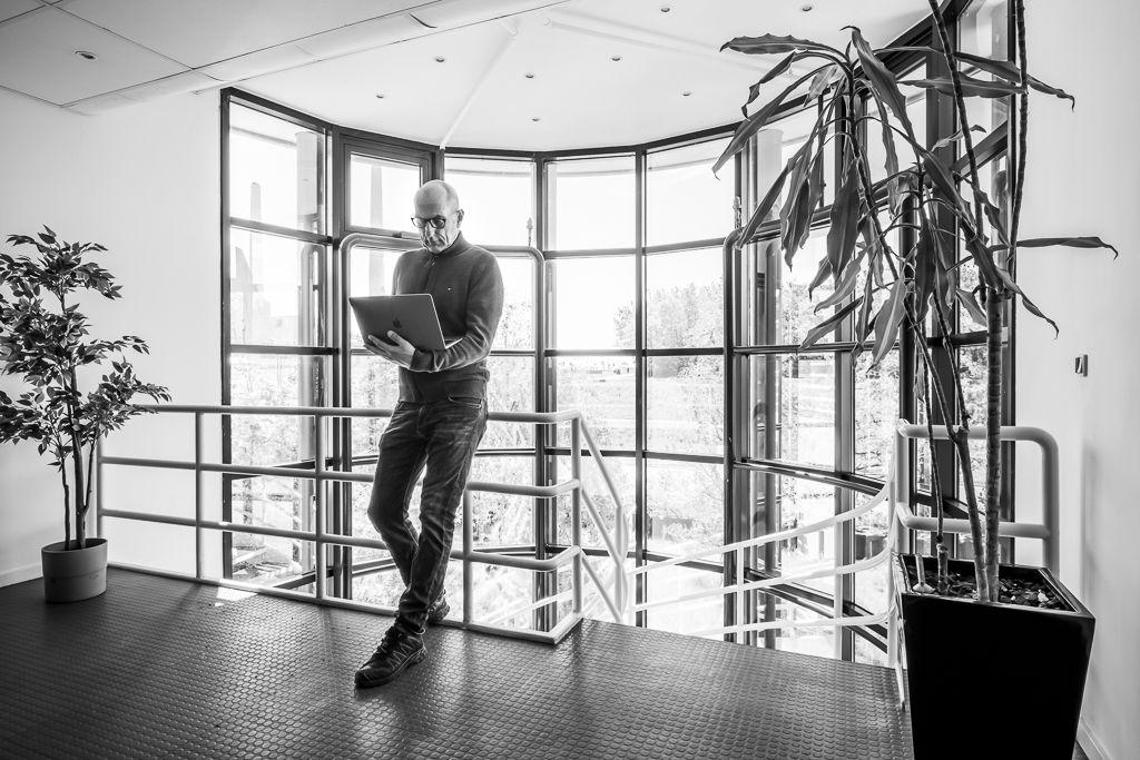 En mand i briller og sko står og læser på en bærbar computer i et moderne kontorbygning.