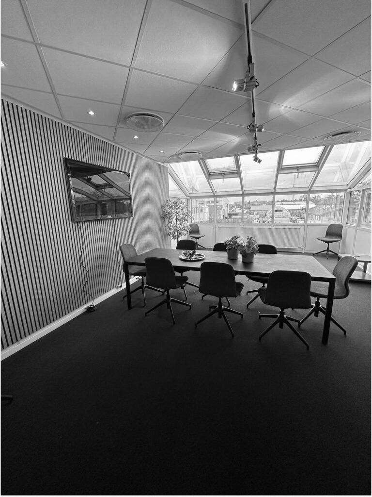 Mødelokalet med lange borde, stole og plantepotter i hjørnet samt en monitor på væggen