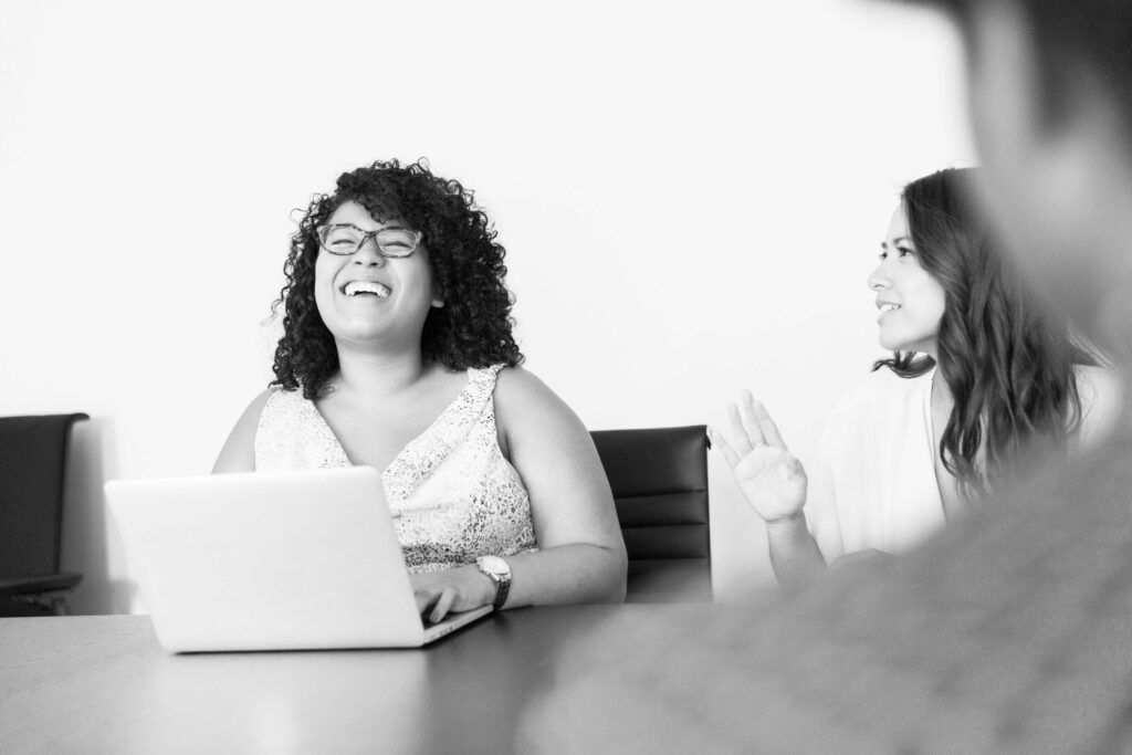 To kvinder sidder ved et bord med en bærbar computer og taler til hinanden. En af dem bærer briller og har en smilende og