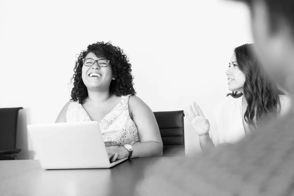 To kvinder sidder ved et bord med en bærbar computer og taler til hinanden. En af dem bærer briller og har en smilende og