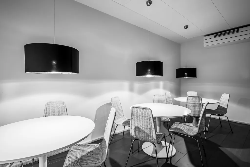 Værelse med fire runde borde og flere stole. Loftet er hvidt, og der er tre store pendant-lamper med sorte kuppelrør. Gulvet