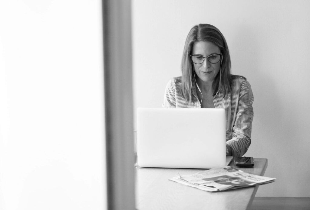 En kvinde sidder og arbejder på en bærbar computer med en tablet og en avis på bordet foran hende