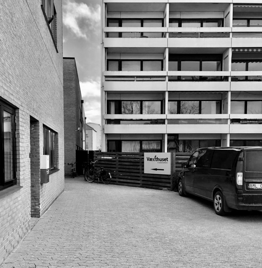 En parkeringsplads med en svart kombiparkeret. Der er flere cykler parkeret ved en træskærm og et skilt med teksten