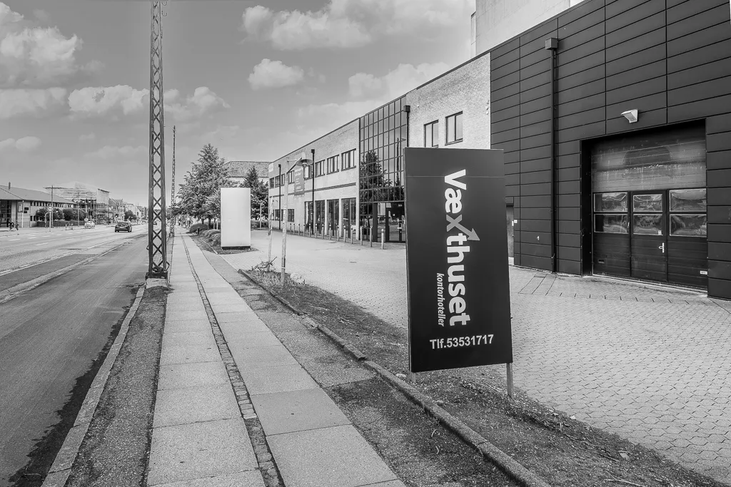 En høj kontorbygning med store ruder og garageport, placeret på hjørnet af en bred gade med en fortov. En stor sort skilt