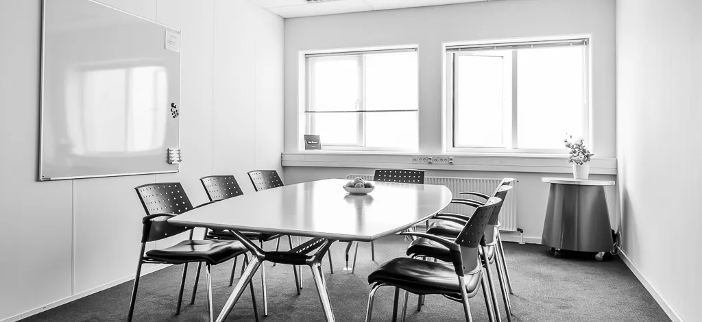 Kontor med bord og stole, tavle og vinduer, hvor der er et bord med frugt og en vase med blomster på.