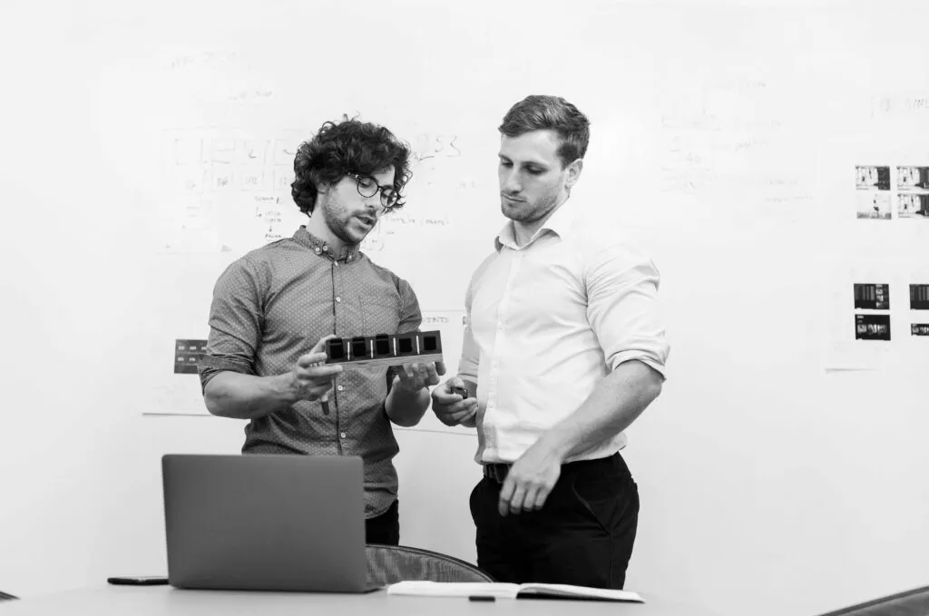 To mænd med briller og skjorte står over en computer og taler om noget og en person har noget at holde i hænderne