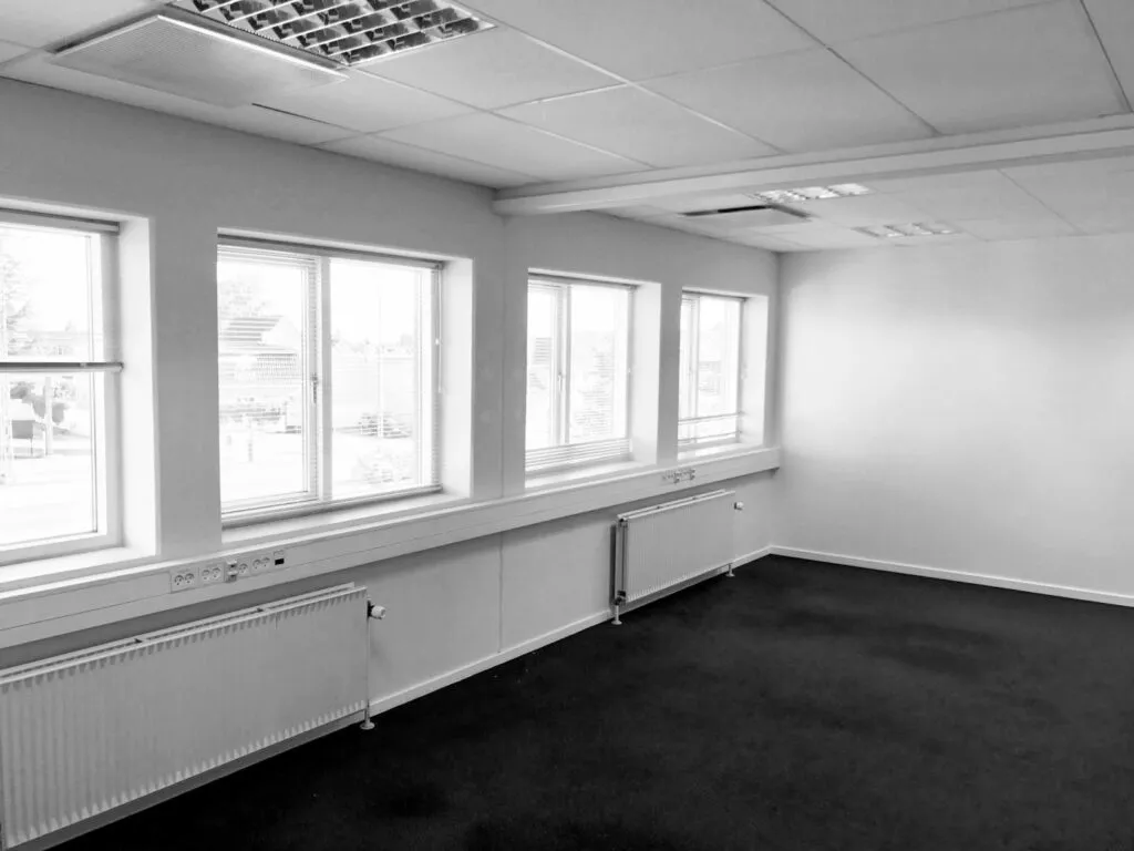 Højloftet kontor med fire vinduer, radiatorer, lyse lofter og en møbleret spiseplads