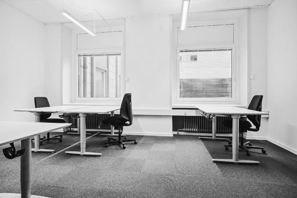 Et tomt kontor med tre borde og fire stole samt to vinduer med gardiner og en radiator under det ene vindue