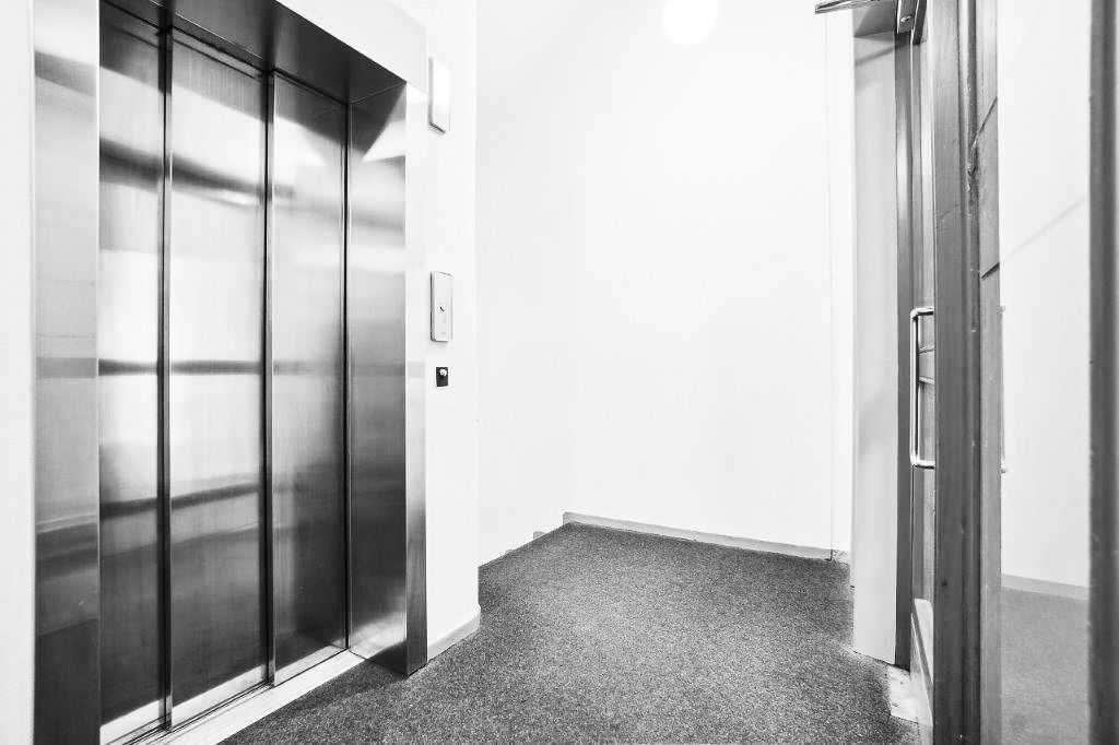 Sort-hvidt billede af et tomt rum med en elevatordør og en lukket vægdør, der fører til et mørkt rum med en lysplet på loftet