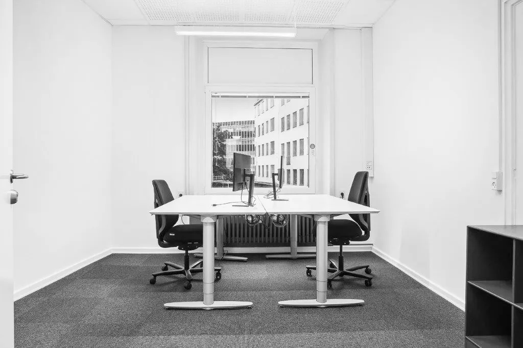 Et kontorlokum med højt til loftet og moderne indretning. Et stort vindue med udsigt til en bygning og et hvidt vægmaleri.