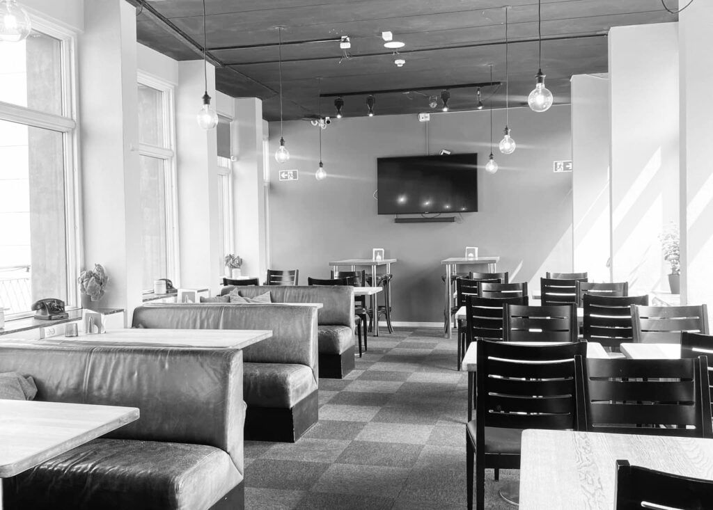 Sort-hvid foto af et tomt café med bord, stole, sofaer og tv-skærm i loftet og plante på vindueskarme