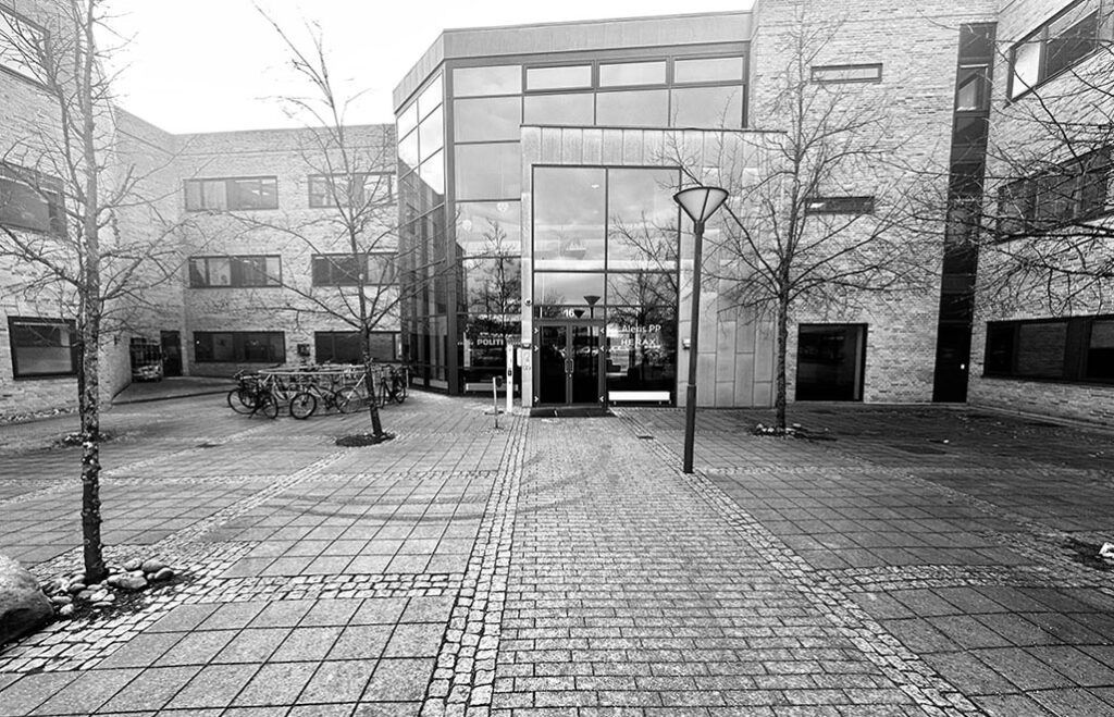 En kontorbygning med glasruder og to indgange, hvor der er flere cykler parkeret udenfor og et søjlelys på højre side