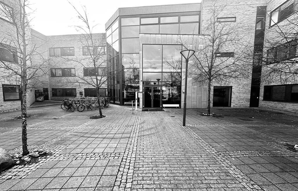 En kontorbygning med glasruder og to indgange, hvor der er flere cykler parkeret udenfor og et søjlelys på højre side