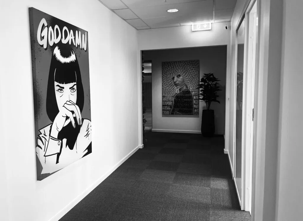 En kontorhvile med en vægmalerier og en plante. En gang med to døre og et lysskilt med pile. Et lyse gulv og et lys på