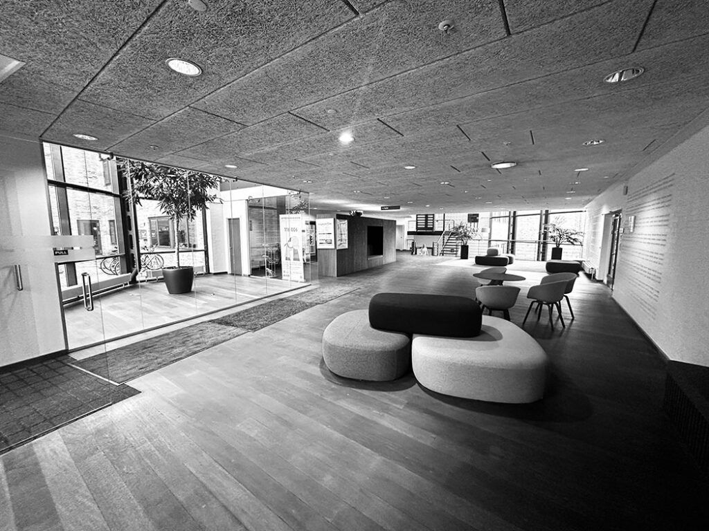 Minimalistisk og moderne lobby med glasvægge, trægulv, og møbler som stole, ottomaner og borde