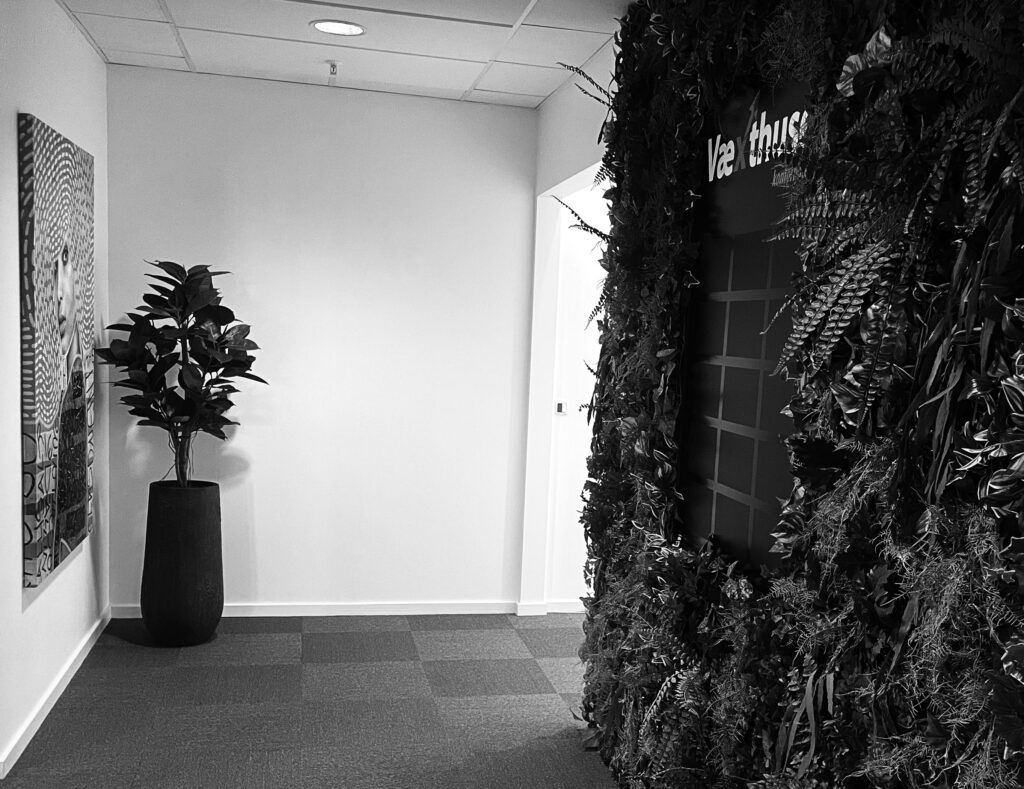 Indvendigt kontorlokaler med gulvtæppe, kunstværk og plante i lysegrå vase, hvid væg og mørk væg med tekst, trægulv og dør