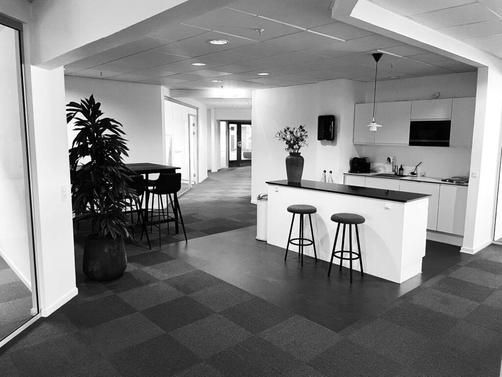 Kontorlokaler med en lyse stue med stolpe, plante og to høje stole samt et køkken med mikrobølgeovn og lyse skabe