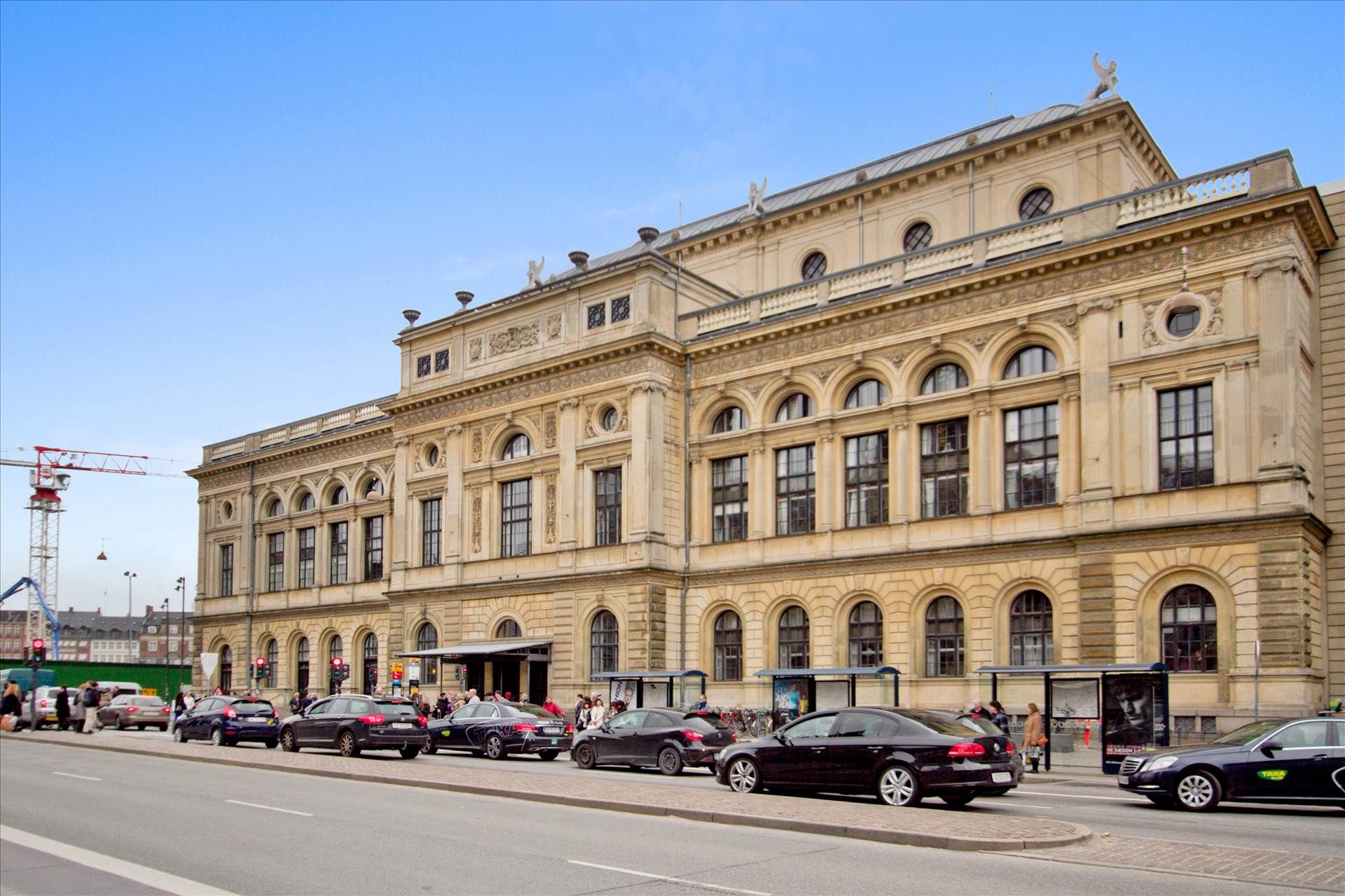 Mange biler parkeret foran bygningen med adgangsskilte og busstop ved siden af, folk går og venter på bussen