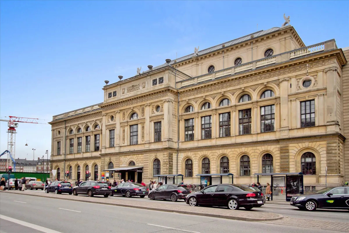 Mange biler parkeret foran bygningen med adgangsskilte og busstop ved siden af, folk går og venter på bussen