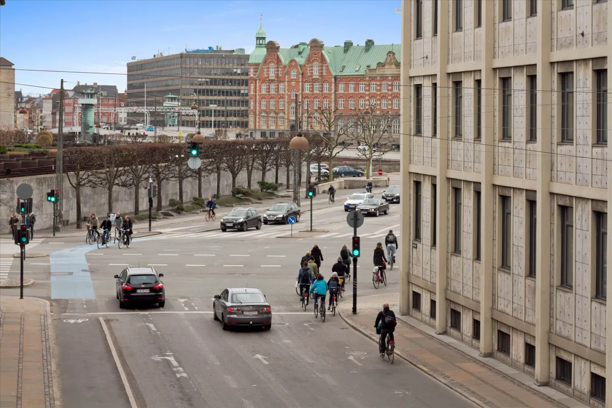 Bygade med cykelstier, biler og folk, store bygninger og træer langs vejen, og rødt og grønt lys på trafiklys
