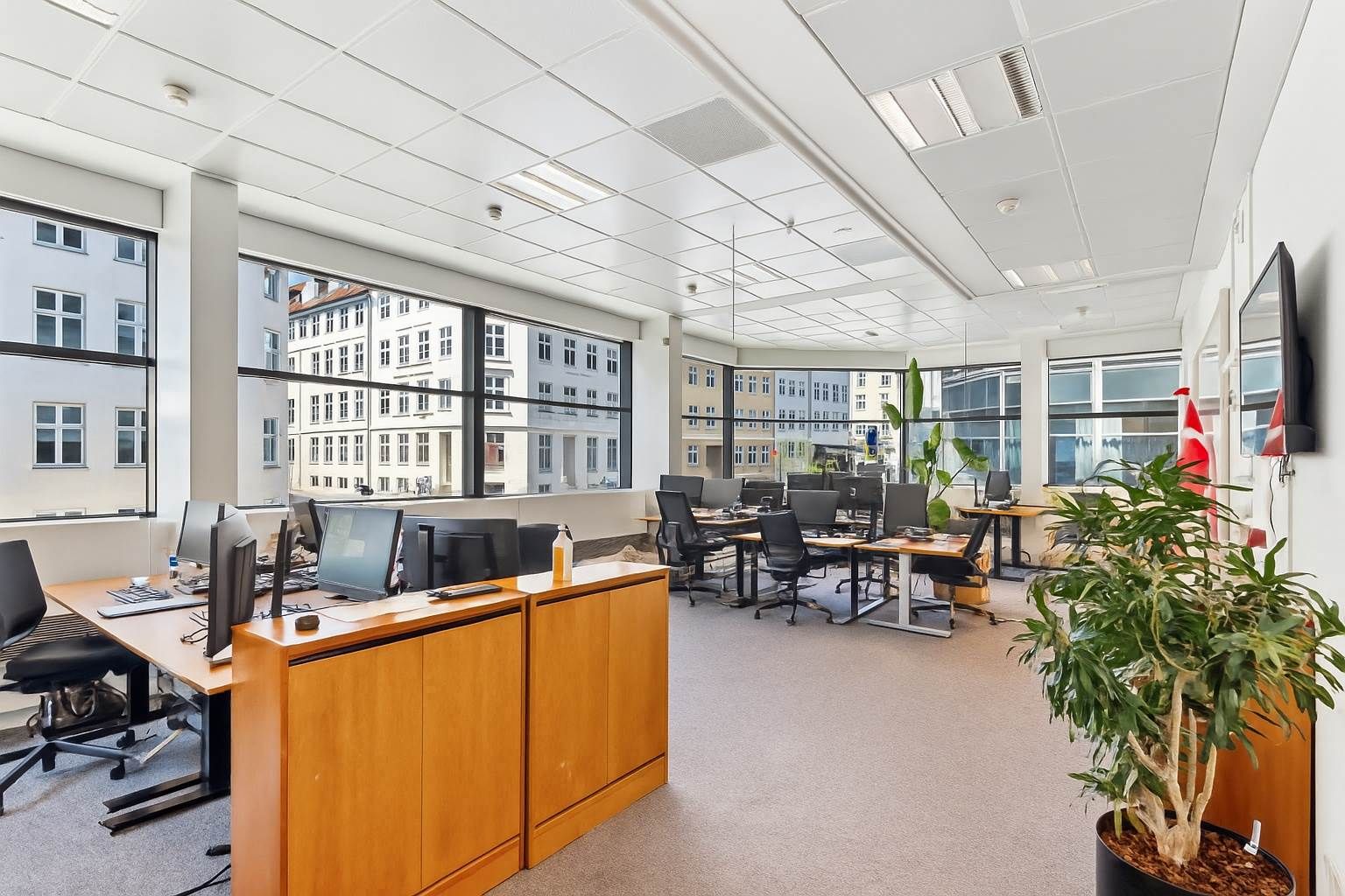 Kontor med gulv til loft vinduer, skriveborde, stolpe, plante og monitorer samt en række tekniske enheder og flag