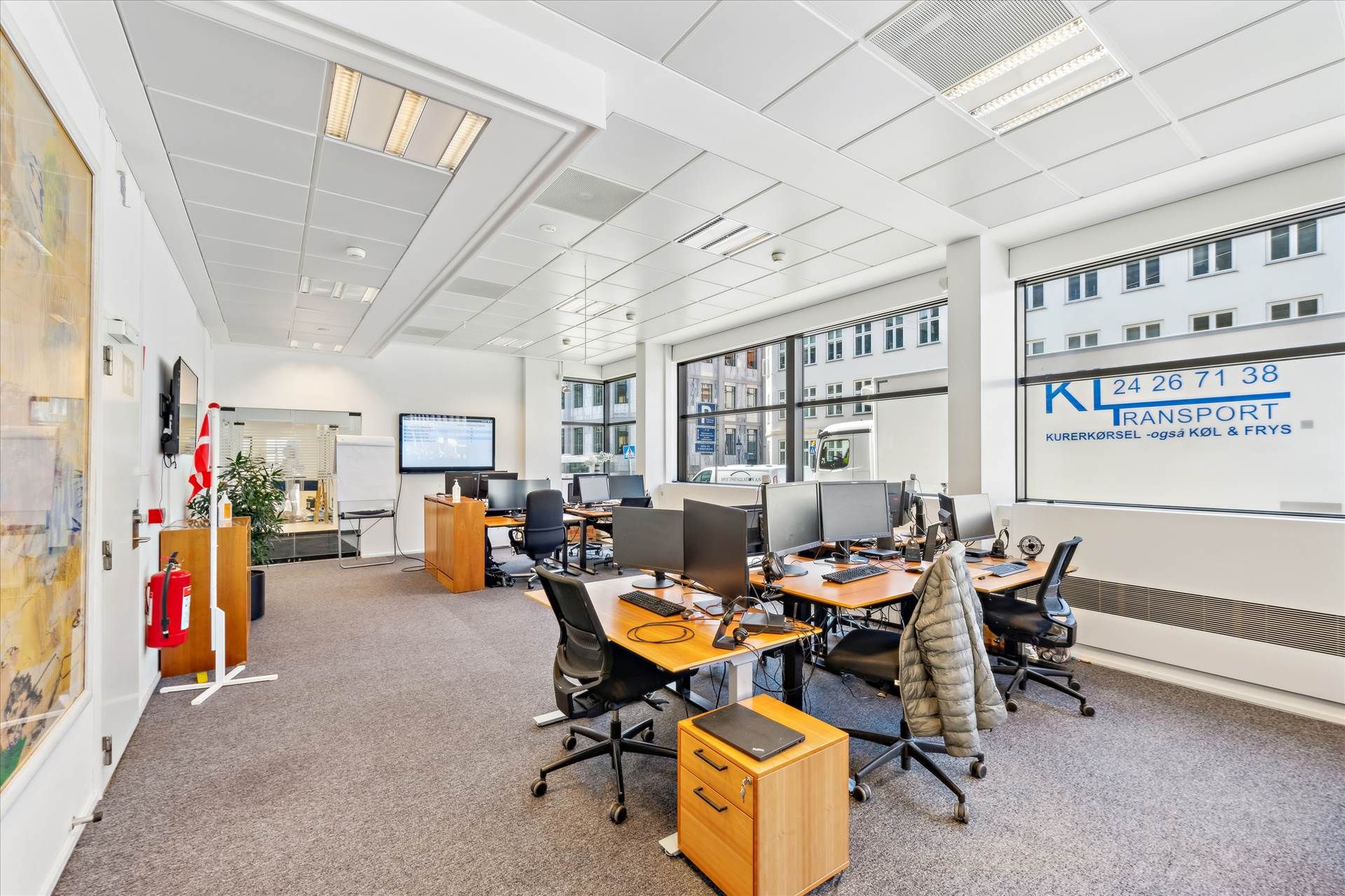 Kontorlokaler med fire skriveborde og computere, lysegule vægge og hvide lofter, vinduer med udsigt til bygninger og veje