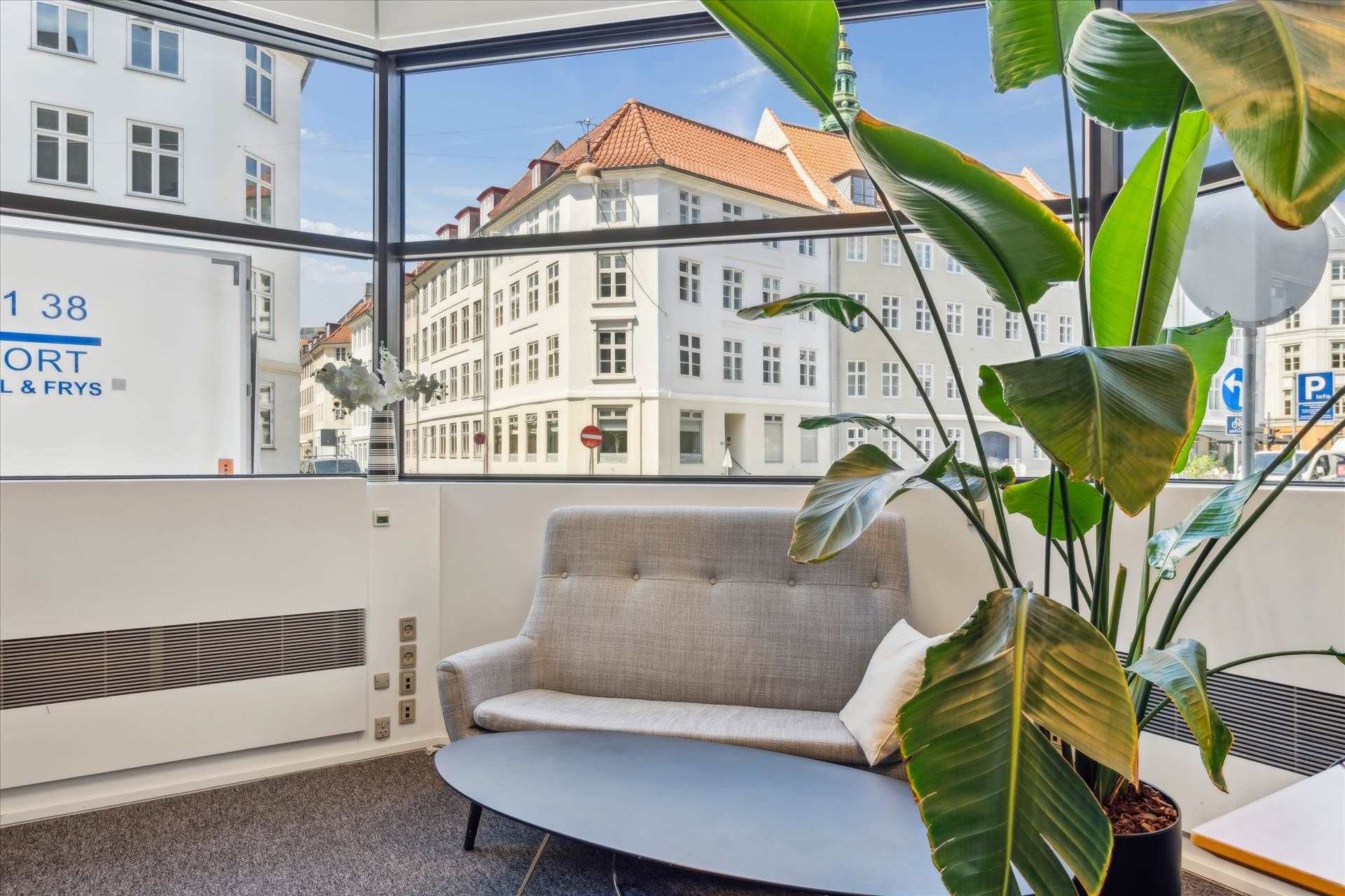 Kontorlokaler med stol, bord, plante og en stor vinduesudsigt til bygning og gadeblik med skilt for Port & Frys