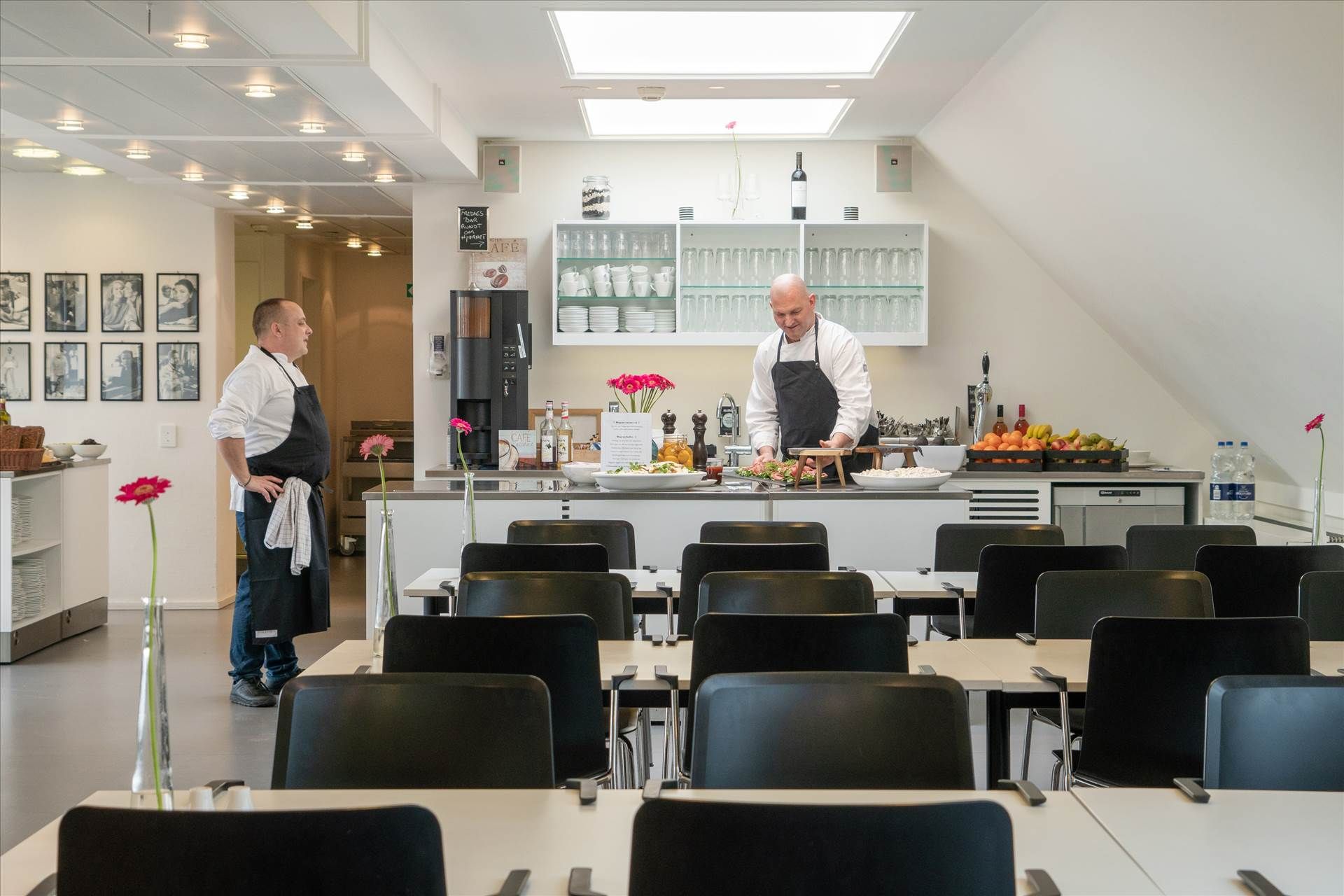 To kokke i sort og hvidt serverer mad til gæster i en spisestue med stole og lange borde