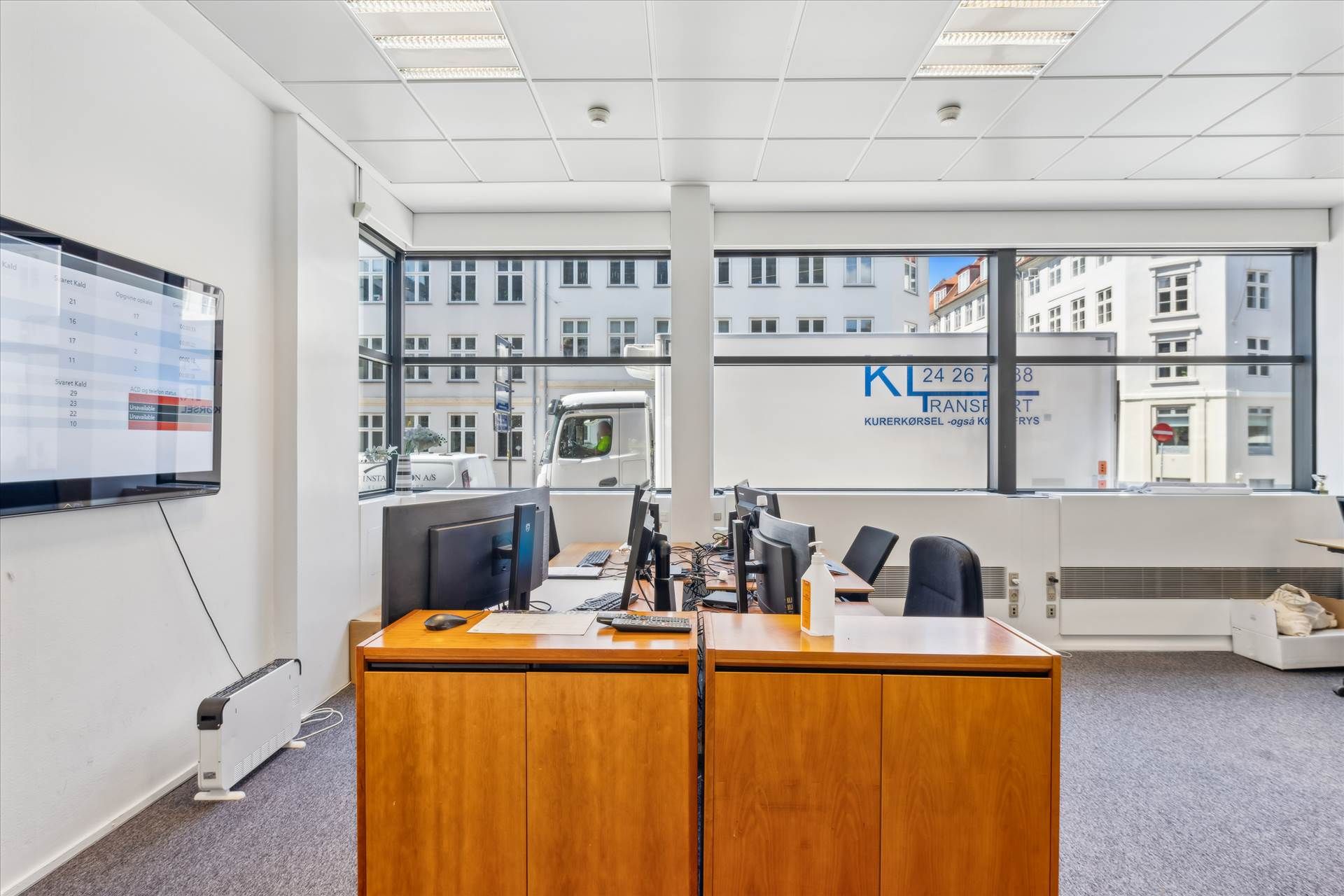 Kontor med computere, skriveborde og monitorer samt glasruder med udsigt til byen og en stående hvid væg med tekst