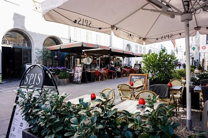 Spis og drikker butik med terrasse og grønne planter. Bord og stole udenfor med parasoller.