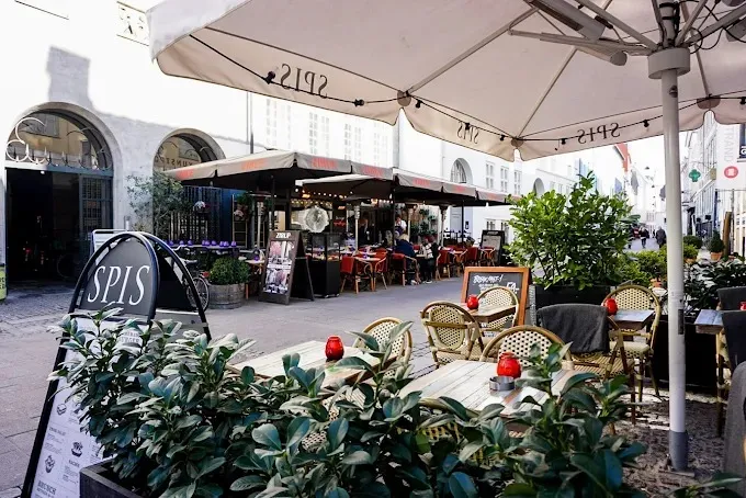 Spis og drikker butik med terrasse og grønne planter. Bord og stole udenfor med parasoller.