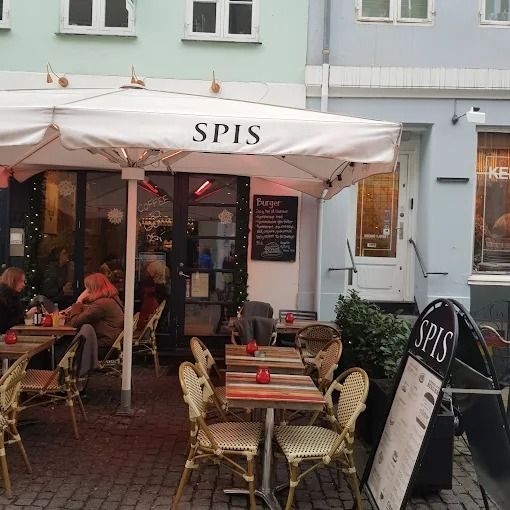 Spis spis restaurant med terrasse, hvor gæster sidder og spiser i en aftensol. På væggen er en skilt med menukort, og det er