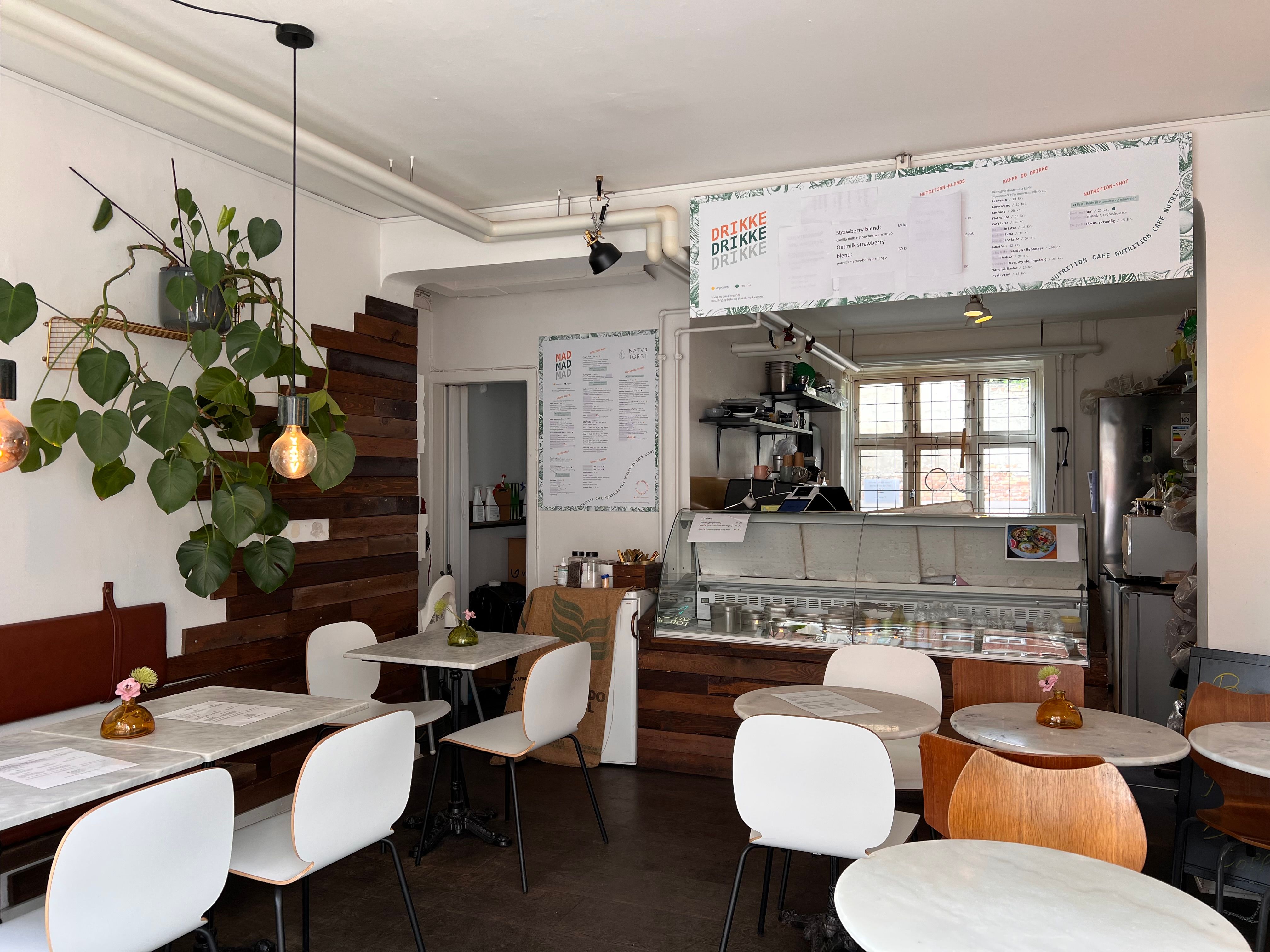 En café med lyse vægge, hvide stole, rundere træborde, loftslampe, plante og menukort med drinks og mad.
