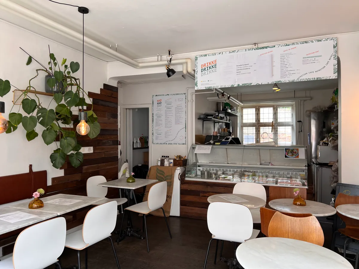 En café med lyse vægge, hvide stole, rundere træborde, loftslampe, plante og menukort med drinks og mad.