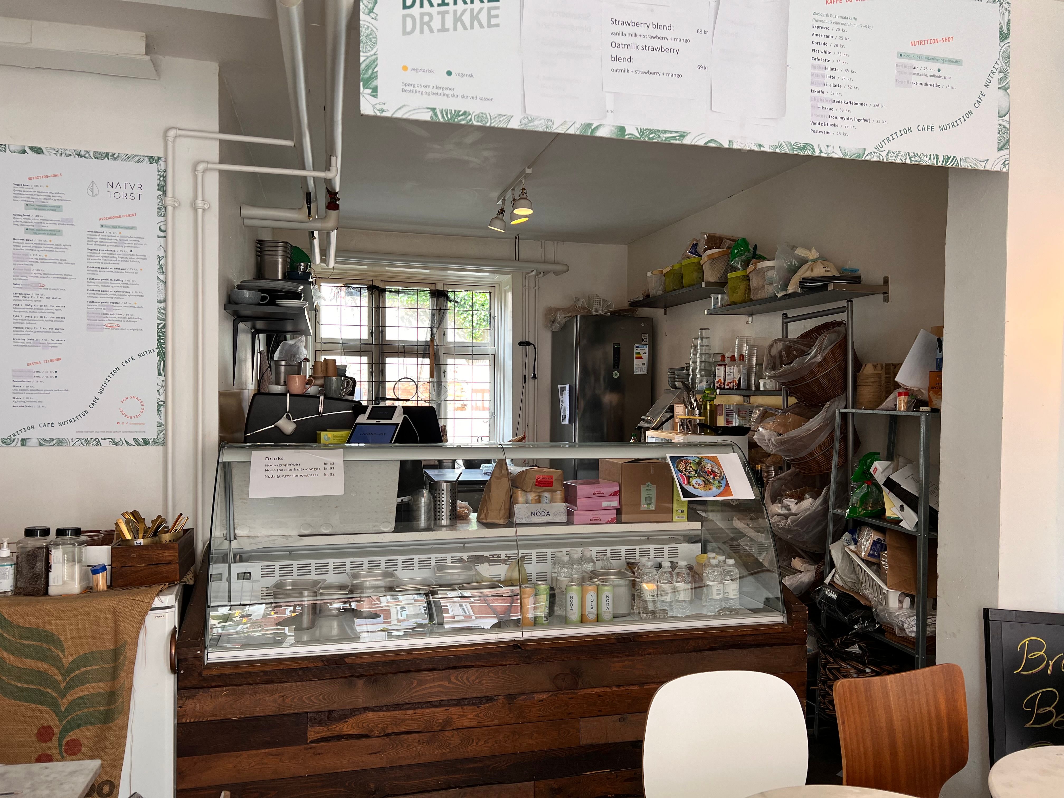 Inde i et cafélokal med et udvalg af drinks og mad i et glasreol, med et menukort og træhylder med produkter