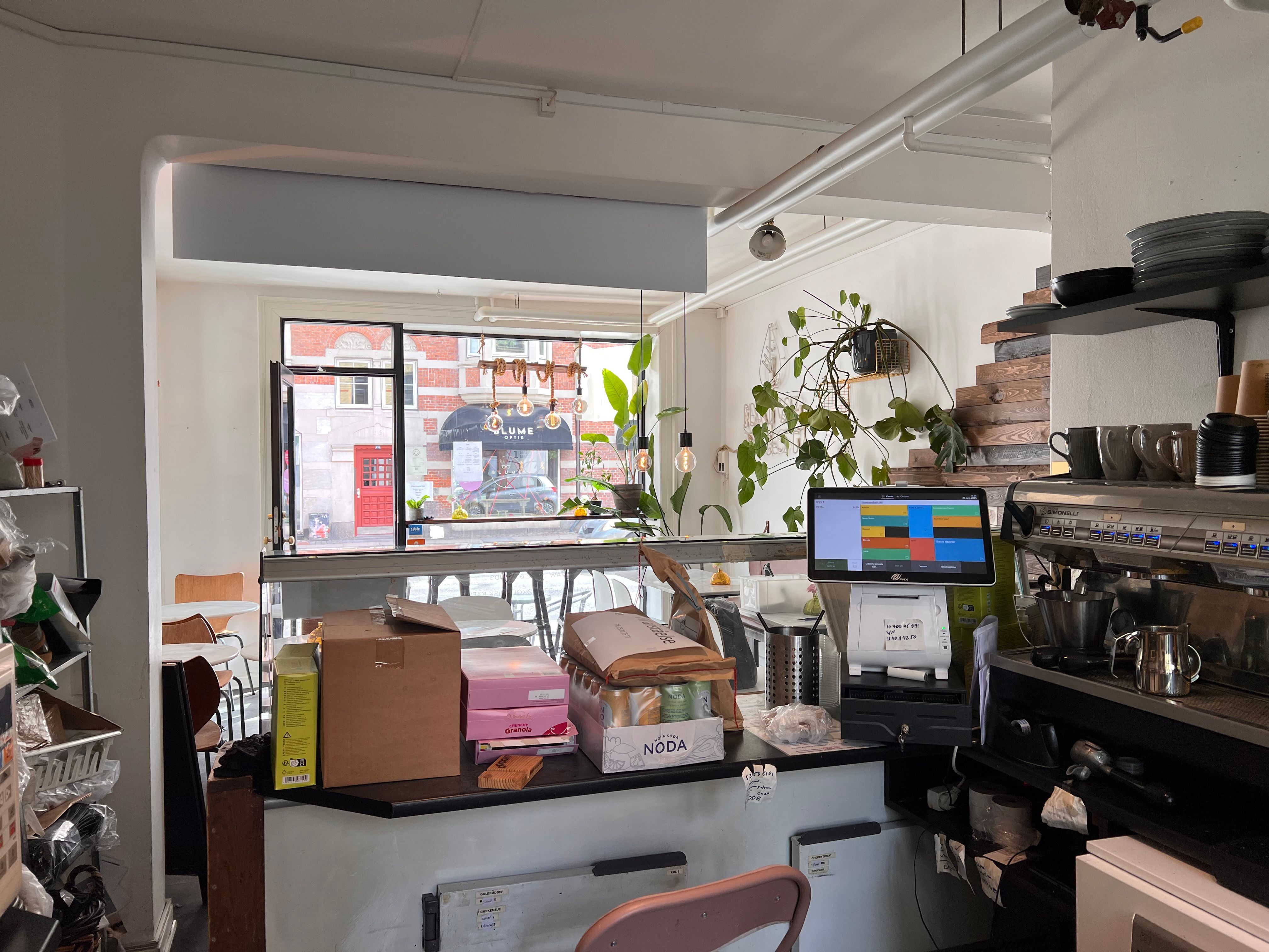 En køkkendel i café med computer, kasseapparat, gryder og kopbeholder samt kaffemaskine og skuffer med varer.