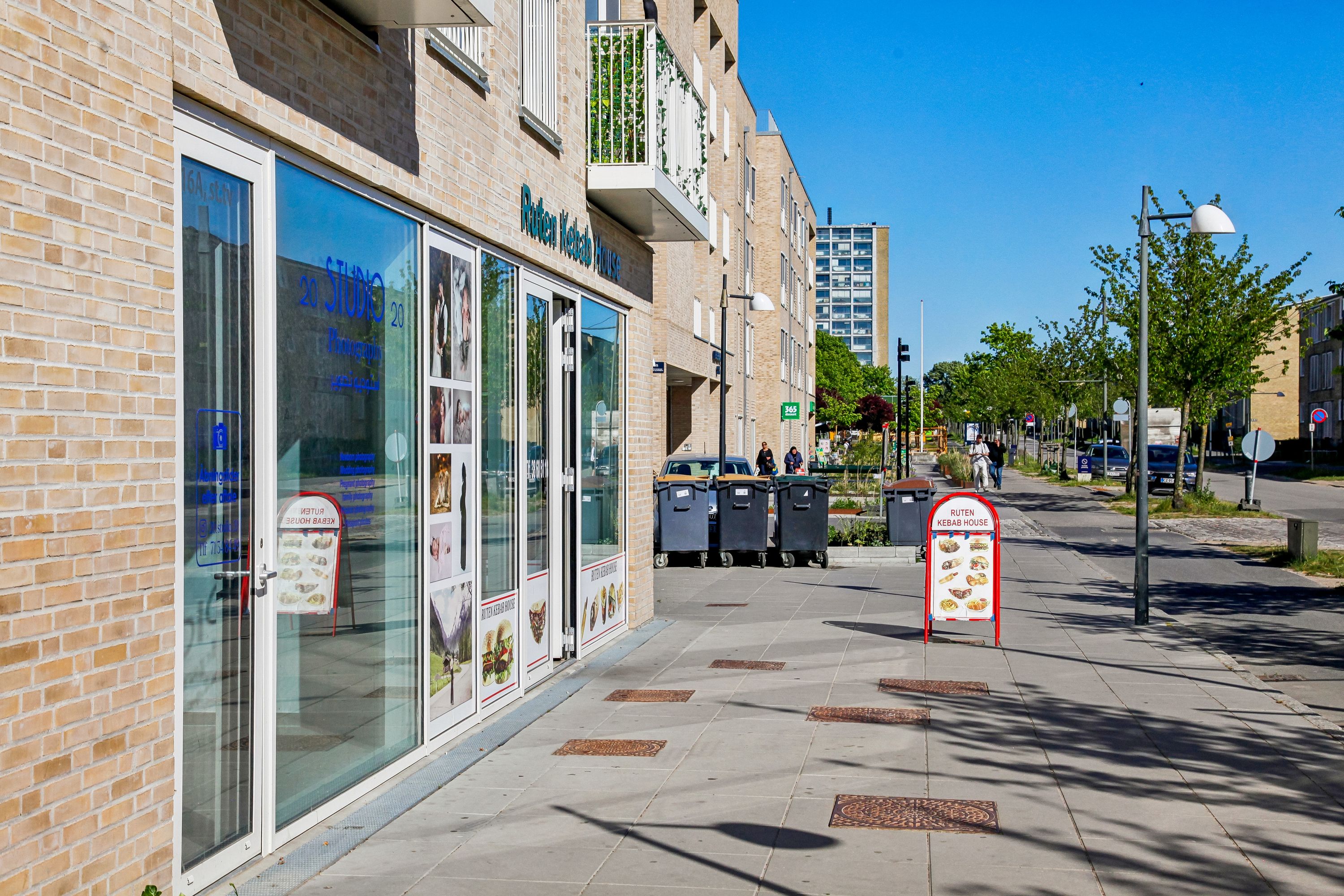 En butik med 20 Studio og Ruten Kebab House i en moderne bygning med grønne buske og parkerede biler på gaden