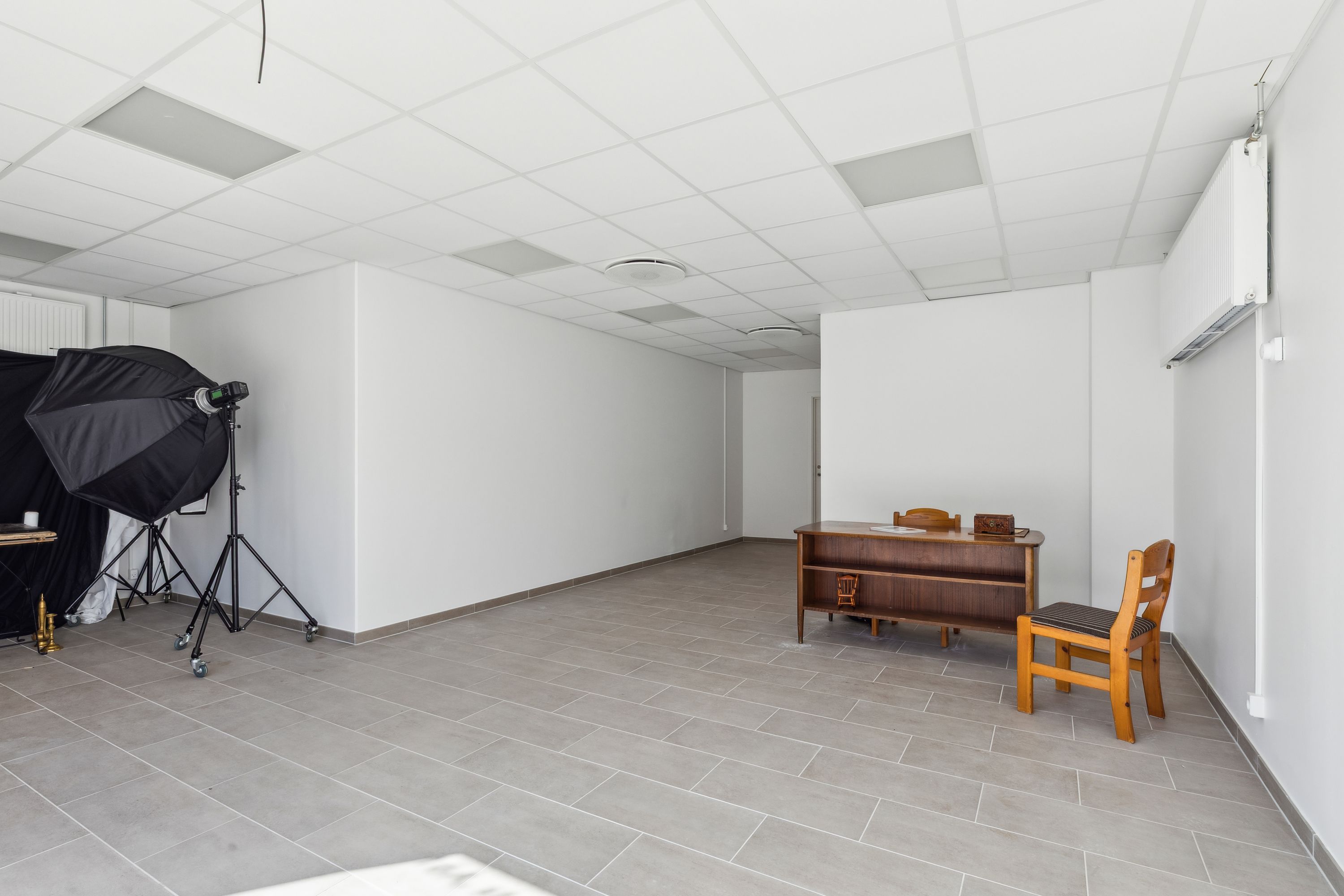 Rum med højt hvide loft og vægge samt gulv med grå flise. Værelset er tomt og har en træskammel og stol samt et stativ med