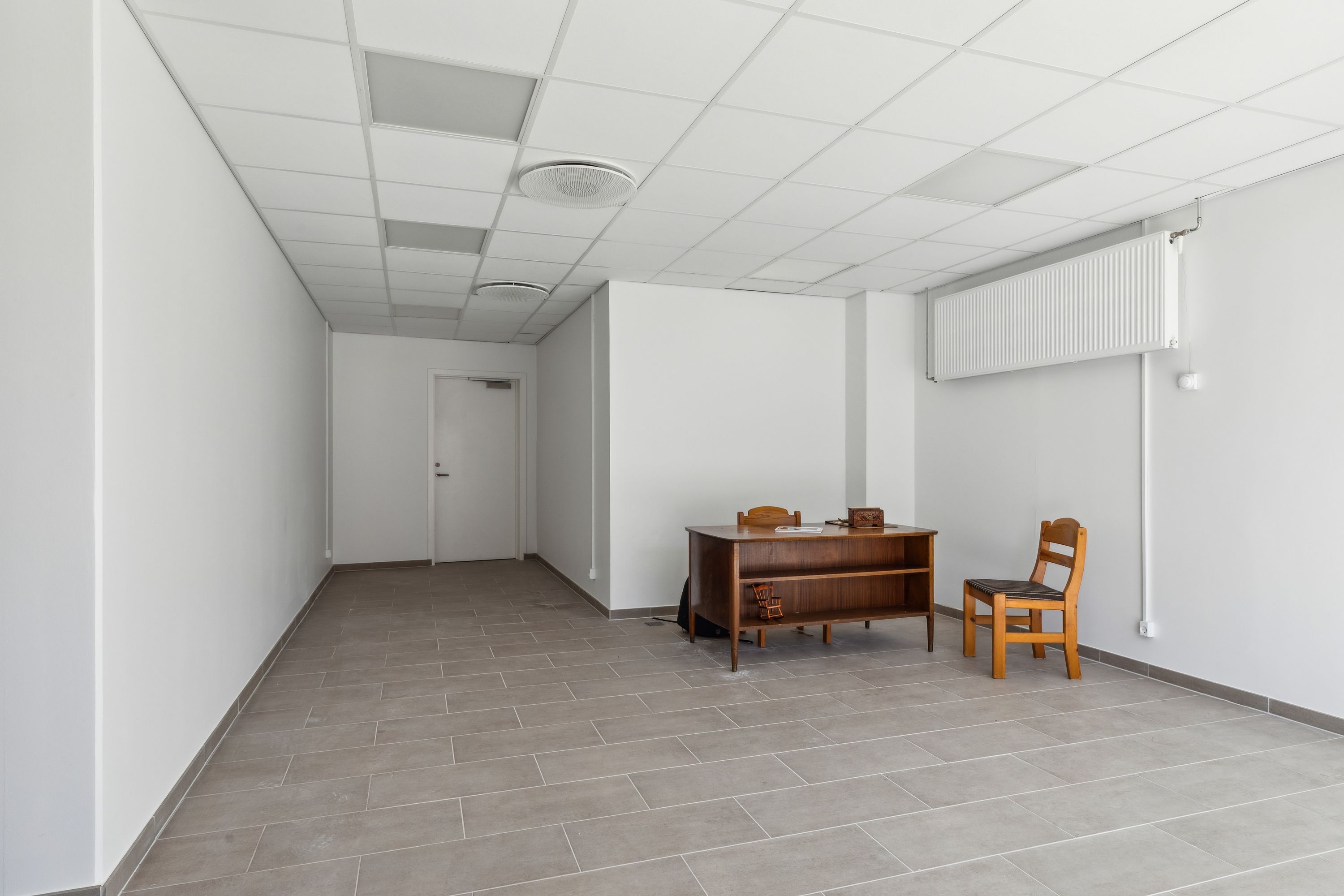 Ledig kontor med lyse vægge, gulv med store kvadratiske fliser og højt loft med hvide paneler. Der er et bord og en stol i