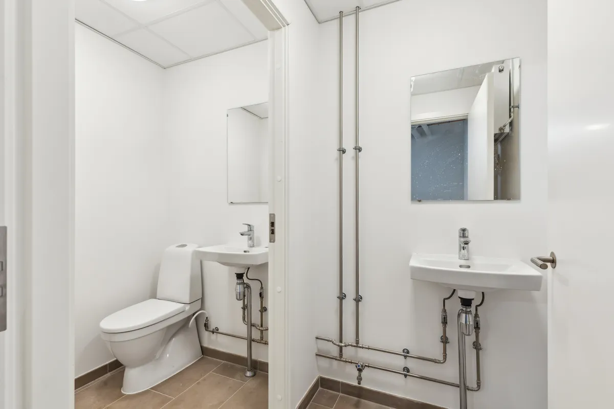 Et lille badeværelse med hvide vægge og loft samt lysegult badeværelsesgulv. Toilet og håndvask med spejl i baggrunden.