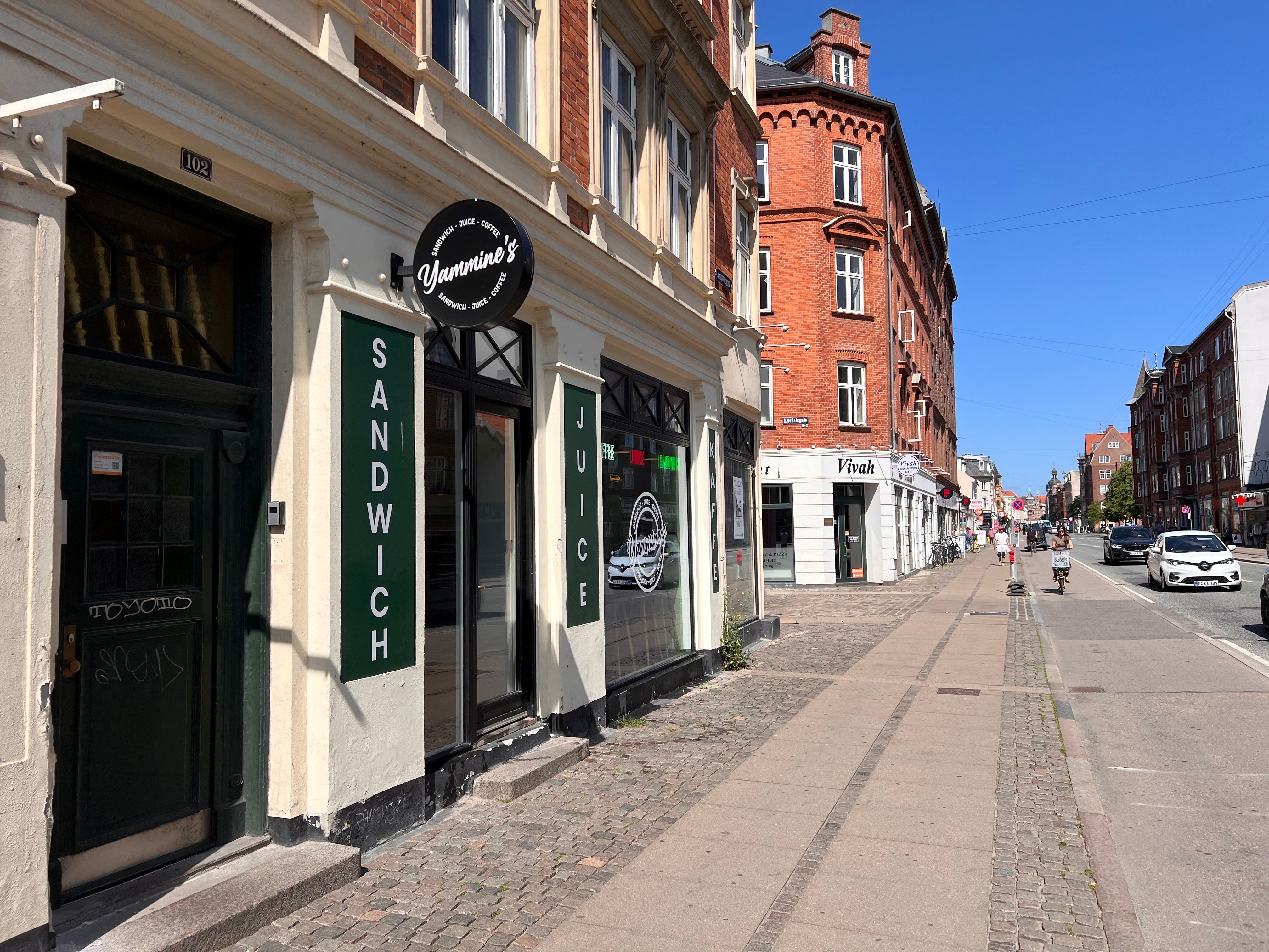 Ymminne's sandwich og juice café med glasfront og indskrift, tæt på andre forretninger og huse, en cyklist og biler på gaden