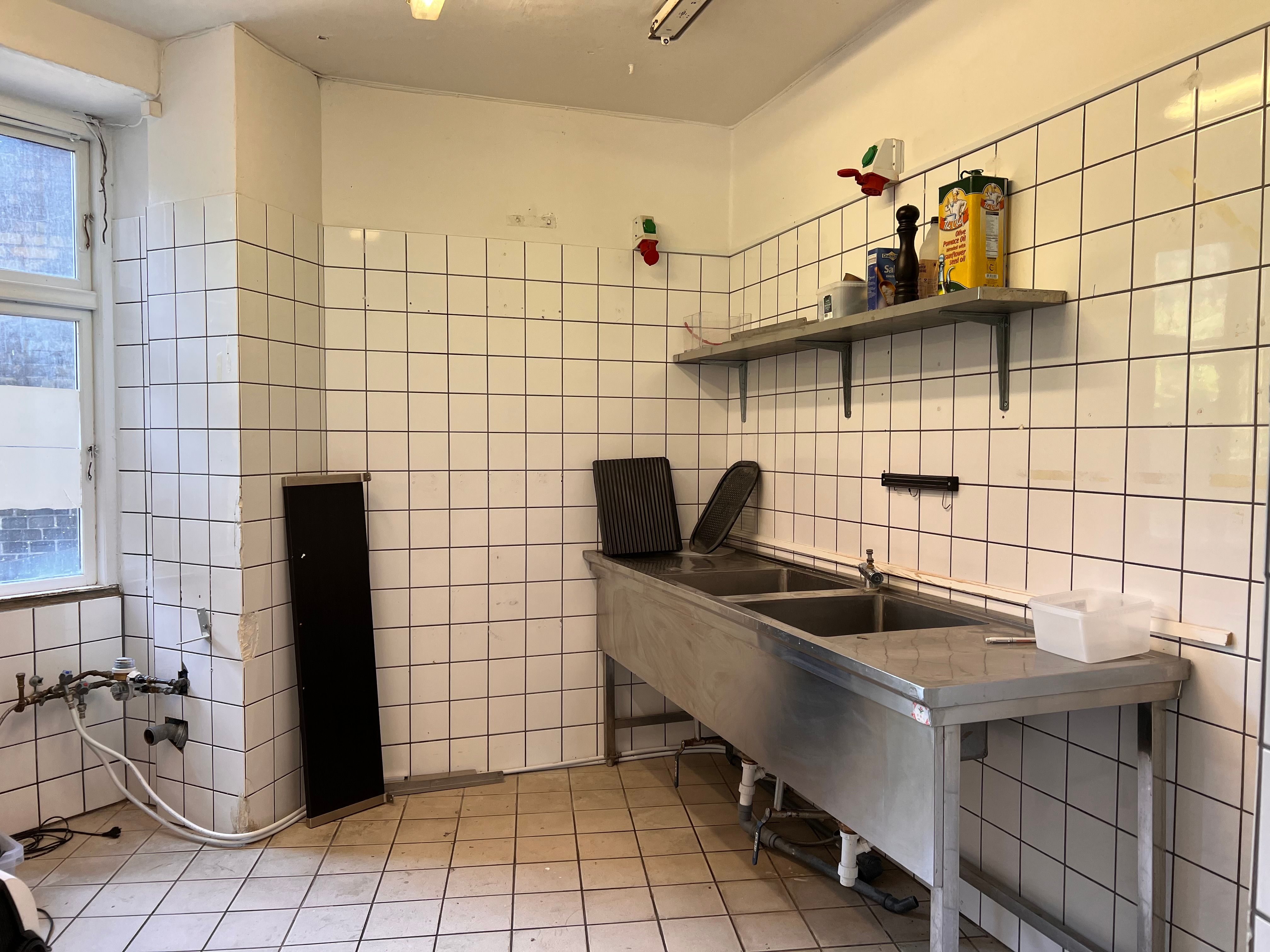 En stor metalarbejdsbord med to vaskebæd og flere beholdere samt to ståhylder med krydderier og andre produkter samt en