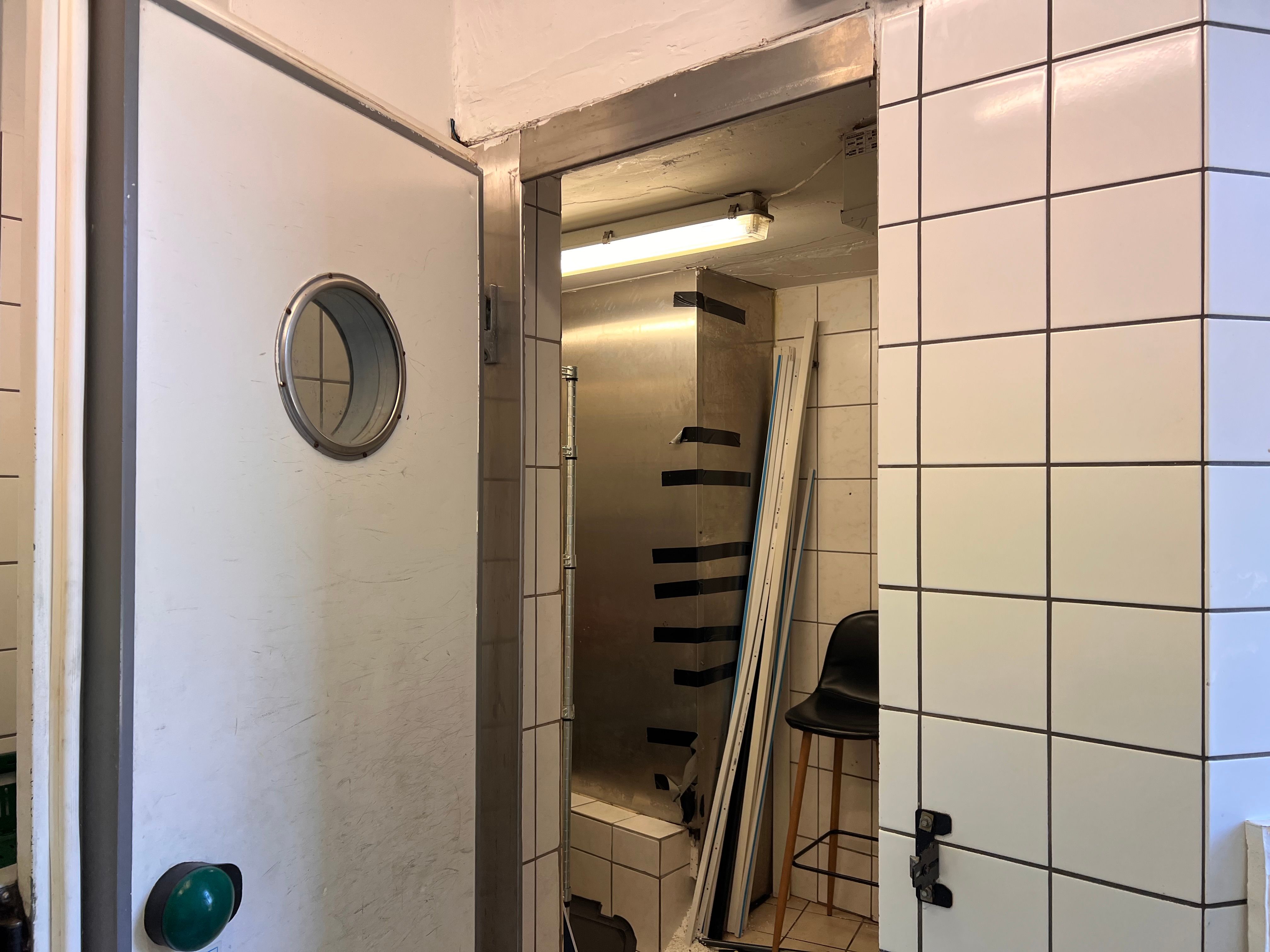 En åben dør og et spejl med en rund udskæring i et hvidt rum med en køkkenskab og en stol i baggrunden