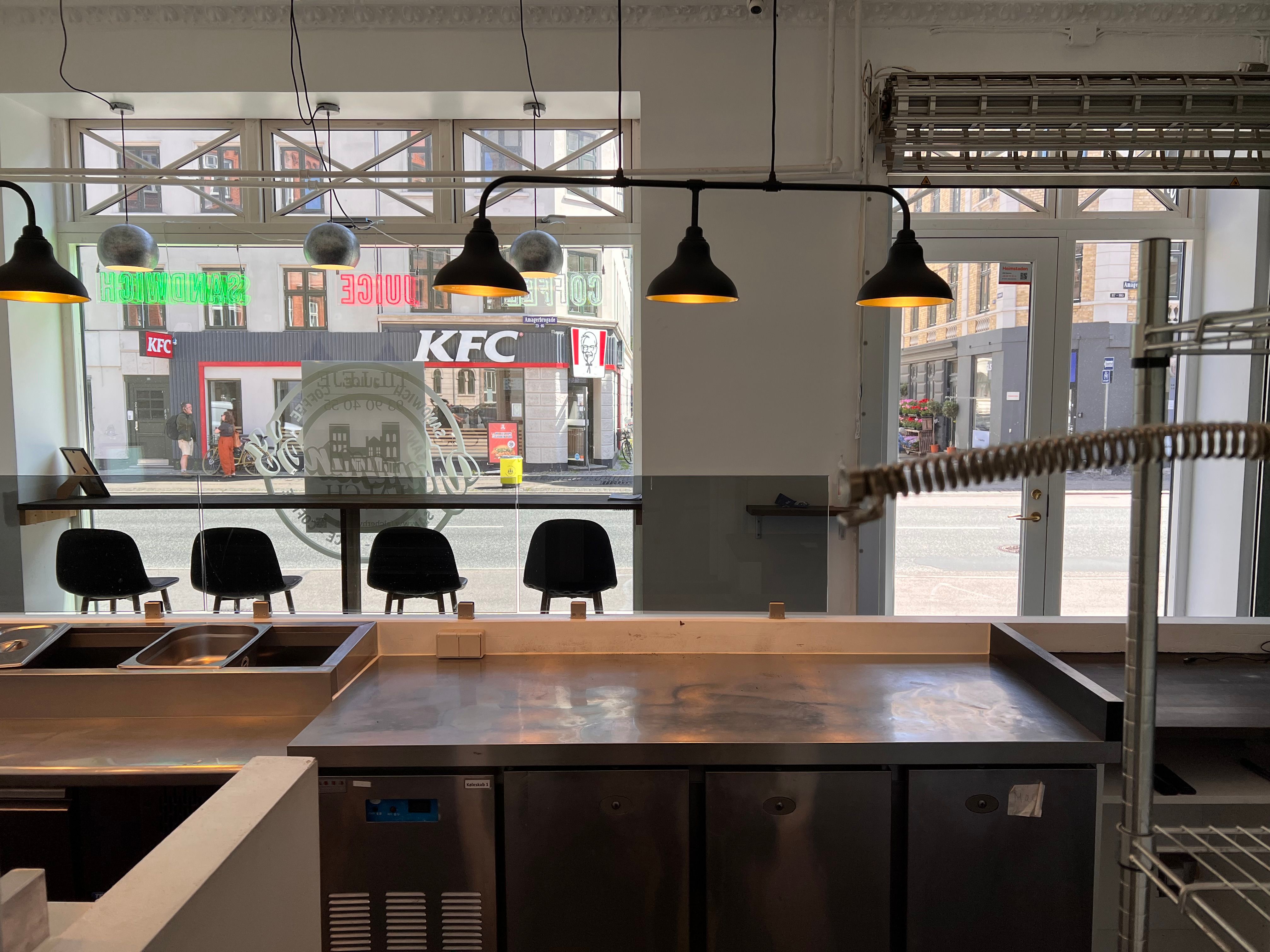 Indvendig af et erhvervslokale med flere køleskabe og en tællere, med fire stole, fire lysekroner og et køkken med en skarp