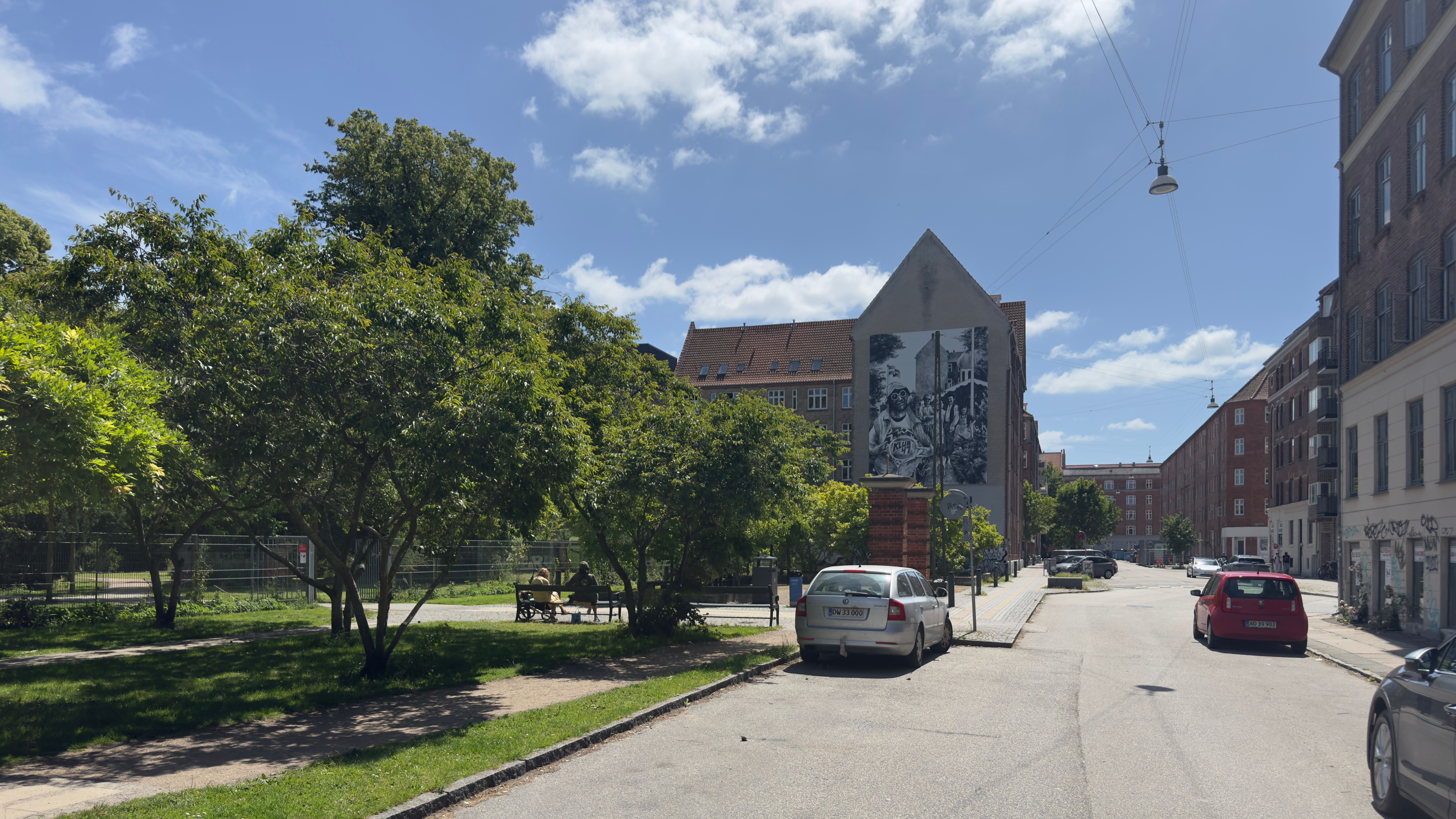 en gade med biler parkeret langs siderne og folk, der går på fortovet, et stort maleri på en bygning og grønne træer og græs